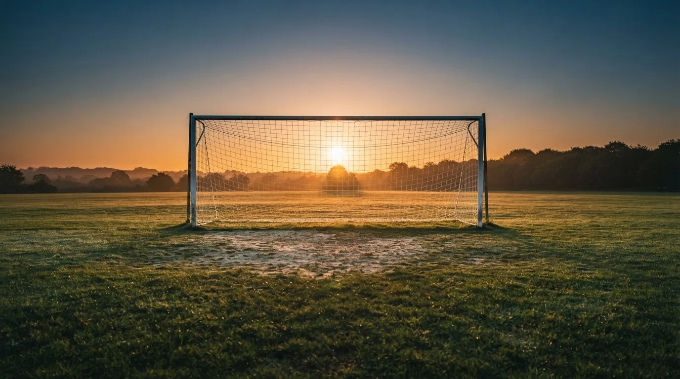 Ein Fußballtor mit Netz bei Sonnenuntergang auf einem leeren Platz, die Schatten der Torpfosten liegen lang auf dem Gras.