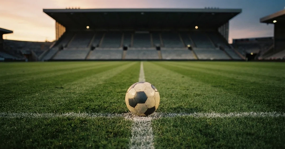 Ein Fußball liegt auf dem Anstoßpunkt eines leeren Rasenplatzes, im Hintergrund eine große Stadiontribüne im Abendlicht.