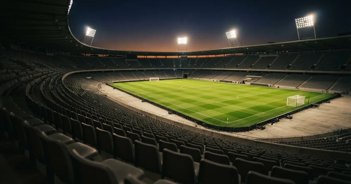 Leere Sitzreihen eines großen Stadions unter Flutlicht, dahinter die ferne Sicht auf den hell beleuchteten Rasen nach Sonnenuntergang.