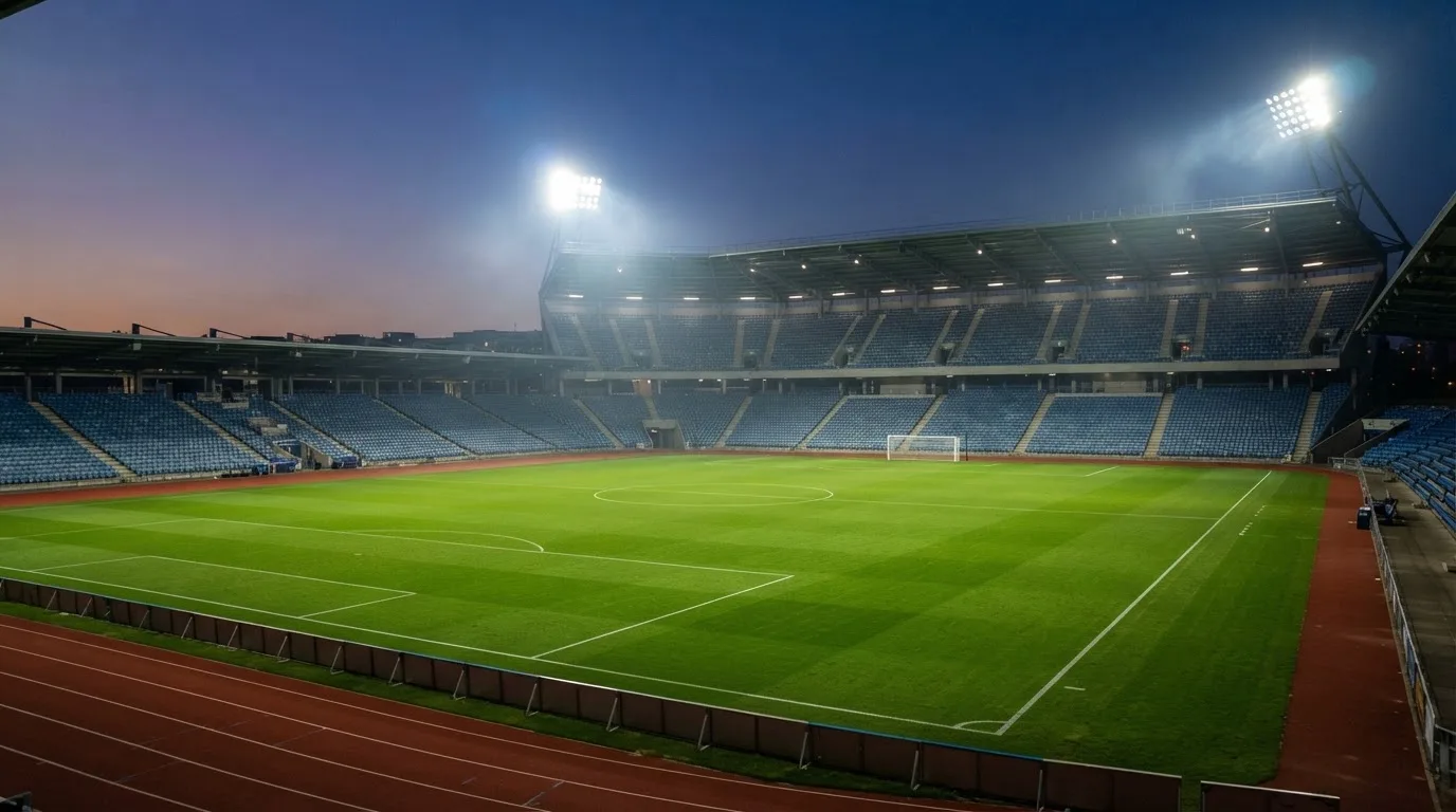 Eine leere Stadiontribüne unter Flutlicht aus der Ferne betrachtet, die Rasenfläche leuchtet grün in der Dämmerung.