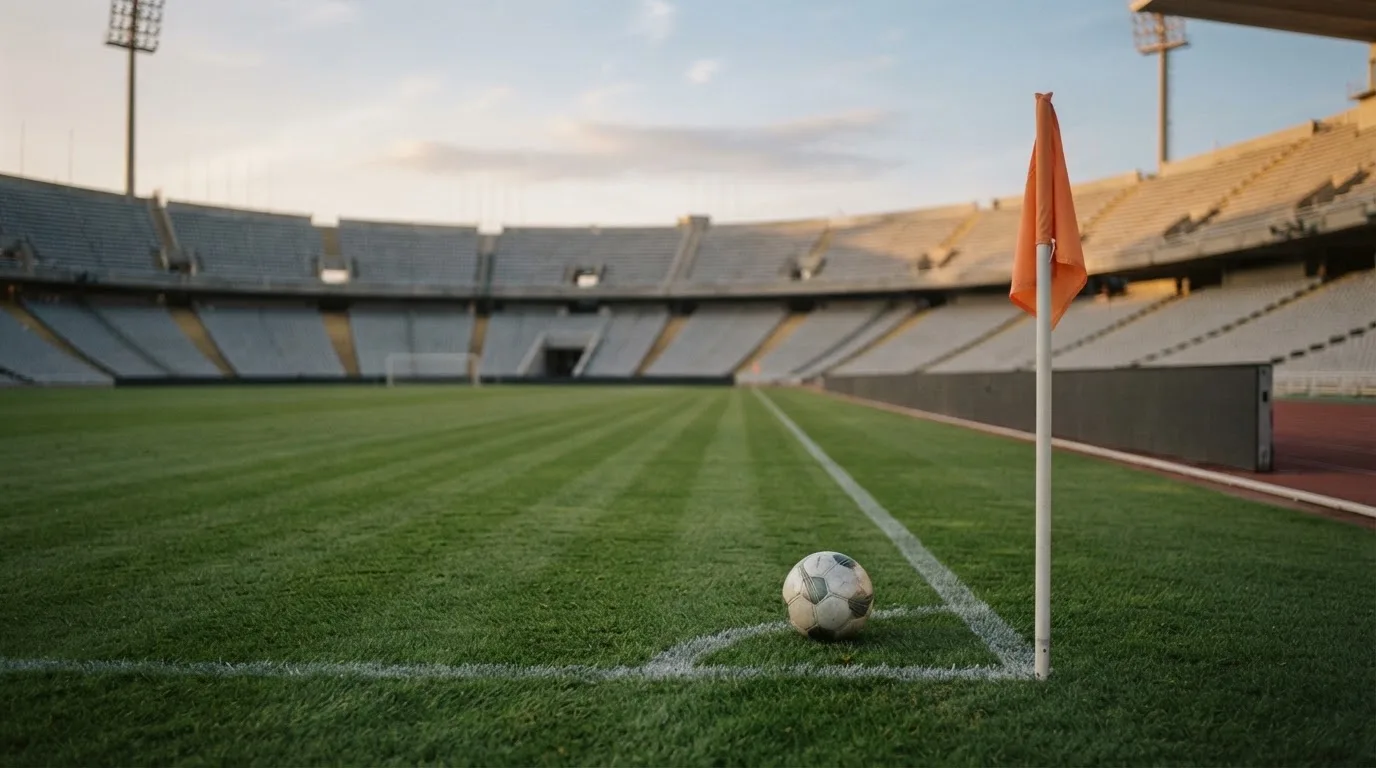 Ein Fußball liegt neben einem Eckfahnen-Pfosten auf einem Rasenplatz, im Hintergrund ein leeres Stadion in der Dämmerung.