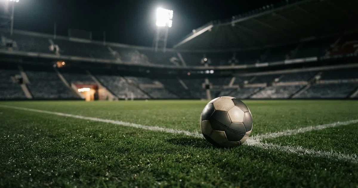 Ein Fußball liegt auf dem Rasen eines leeren Stadions bei Flutlicht, im Hintergrund unscharfe Tribünen ohne erkennbare Beschriftung.