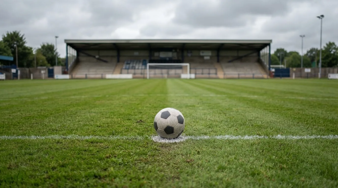 Ein Fußball liegt auf dem Anstoßpunkt eines leeren Rasenplatzes, im Hintergrund eine ruhige Tribüne unter bedecktem Himmel.