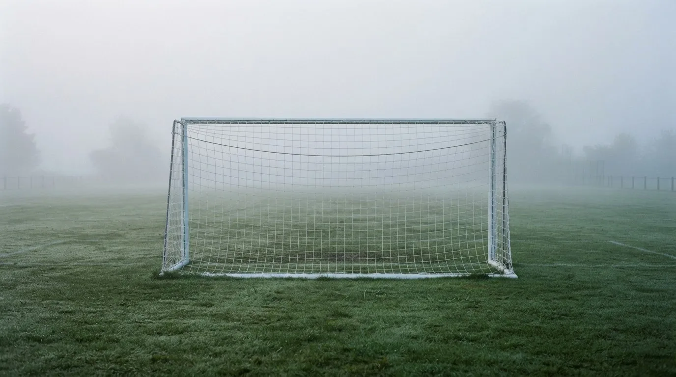 Ein Fußballtor mit Netz im Morgennebel auf einem leeren Platz, die Linien nur schwach zu erkennen.