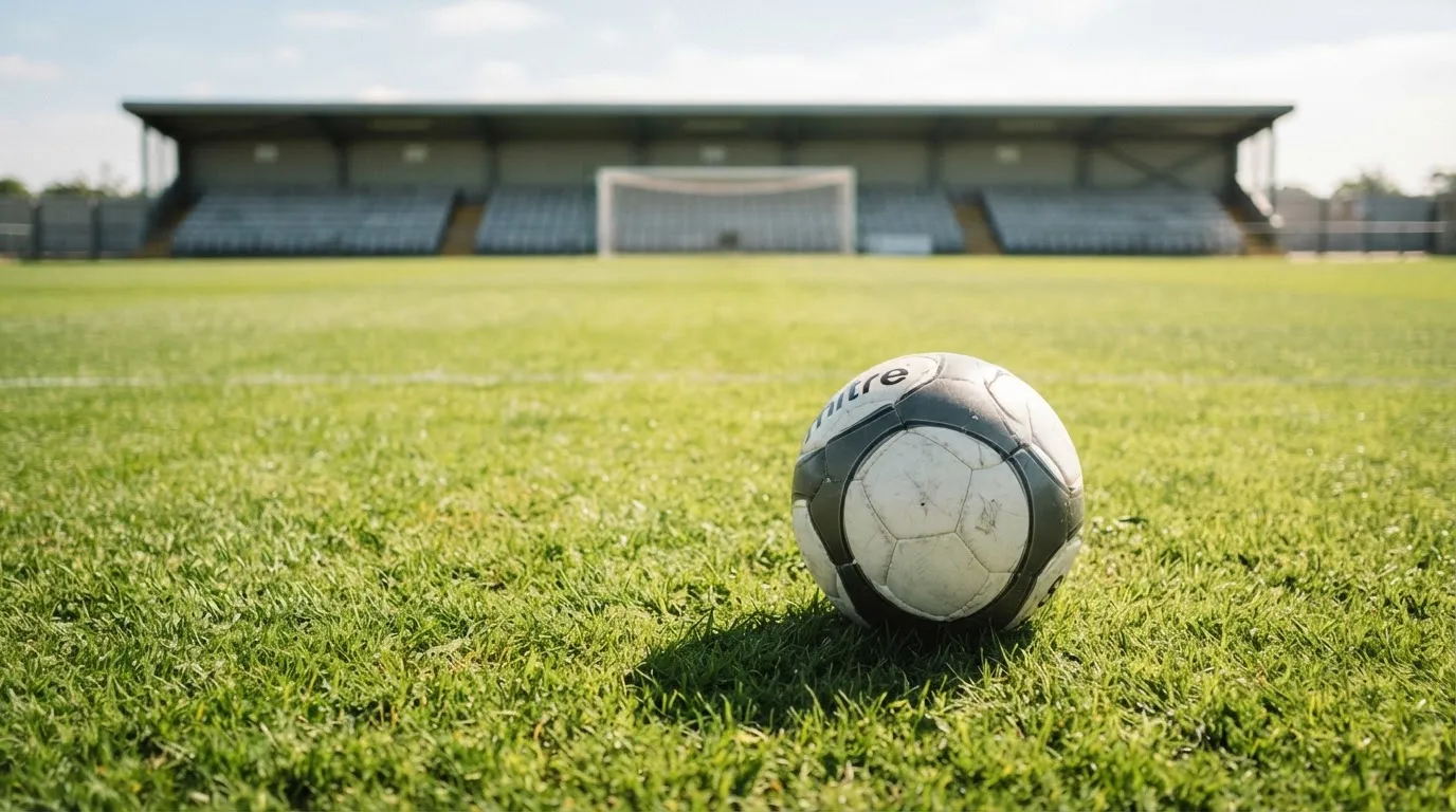 Ein Fußball liegt am Spielfeldrand eines Rasenplatzes, im Hintergrund eine leere Tribüne unter hellem Himmel.