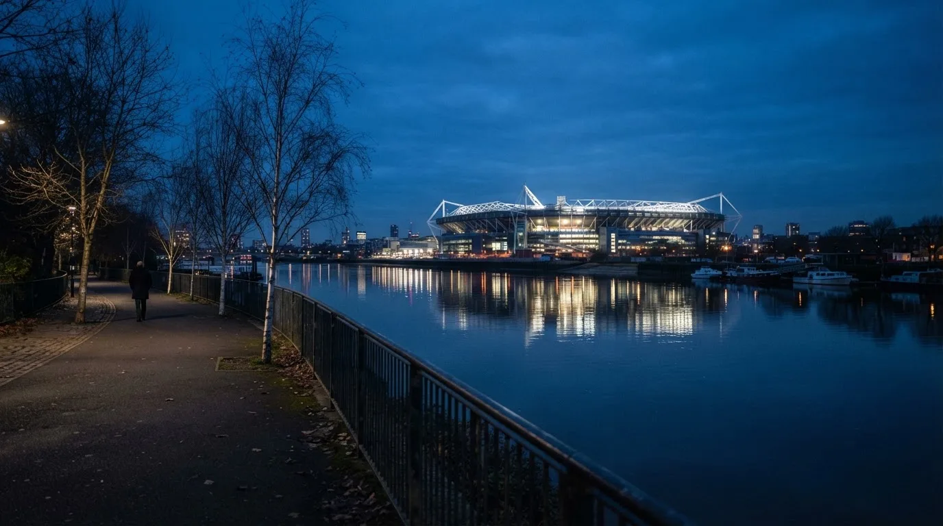 Ein Stadion unter Flutlicht aus der Ferne gesehen, im Vordergrund ein ruhiger Fluss und ein Fußweg.