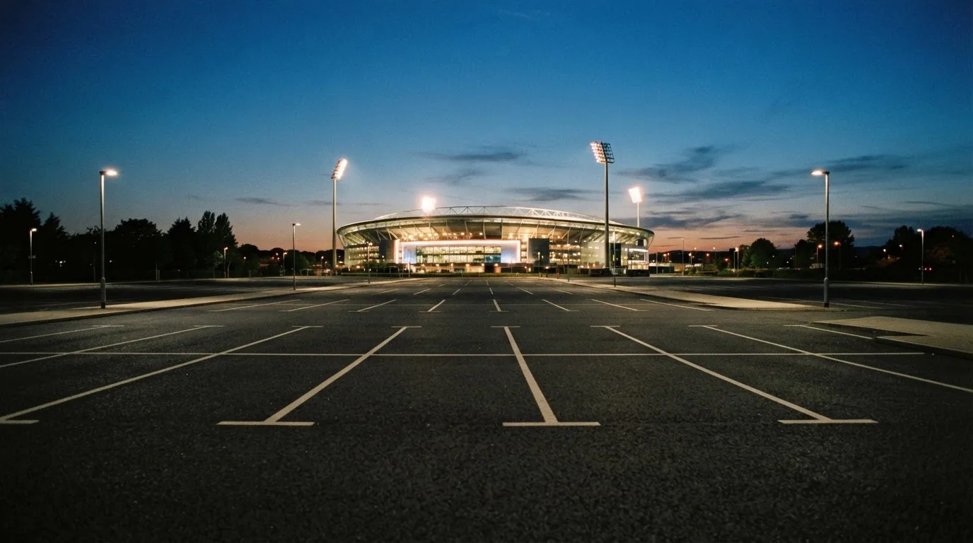 Ein Stadion unter Flutlicht aus der Ferne gesehen, davor ein leerer Parkplatz in der Dämmerung.