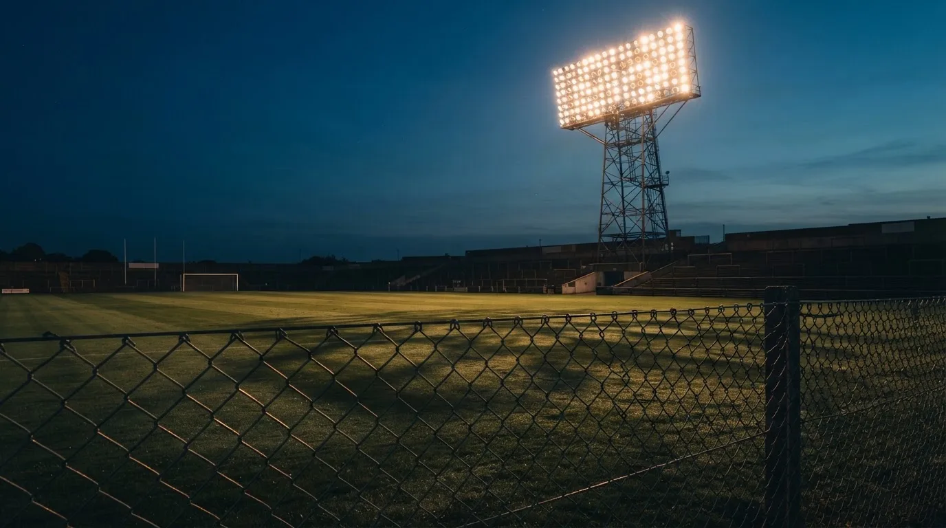 Eine Flutlichtanlage über einem leeren Sportplatz am Abend, im Vordergrund ein Maschendrahtzaun.