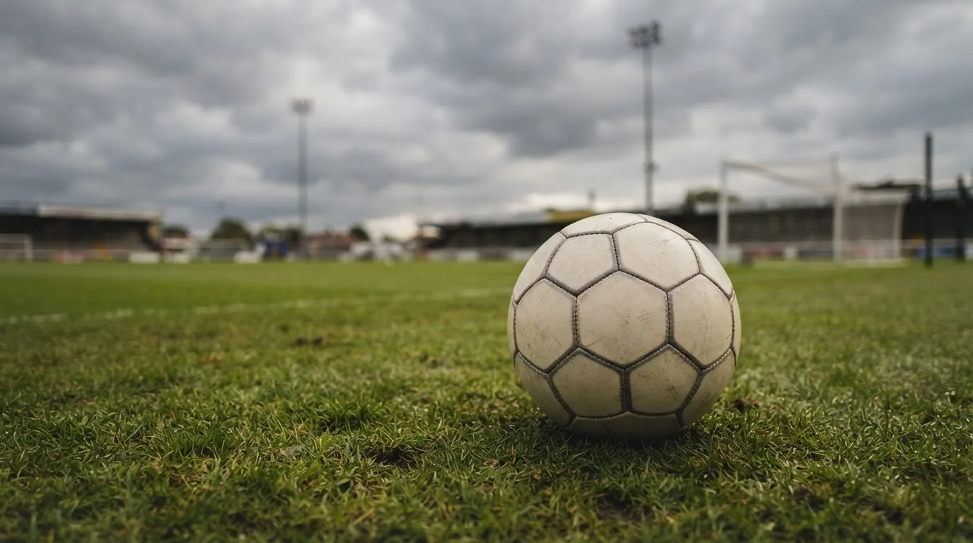 Ein Fußball auf einem Rasenplatz, im Hintergrund Flutlichtmasten und ein bewölkter Himmel.
