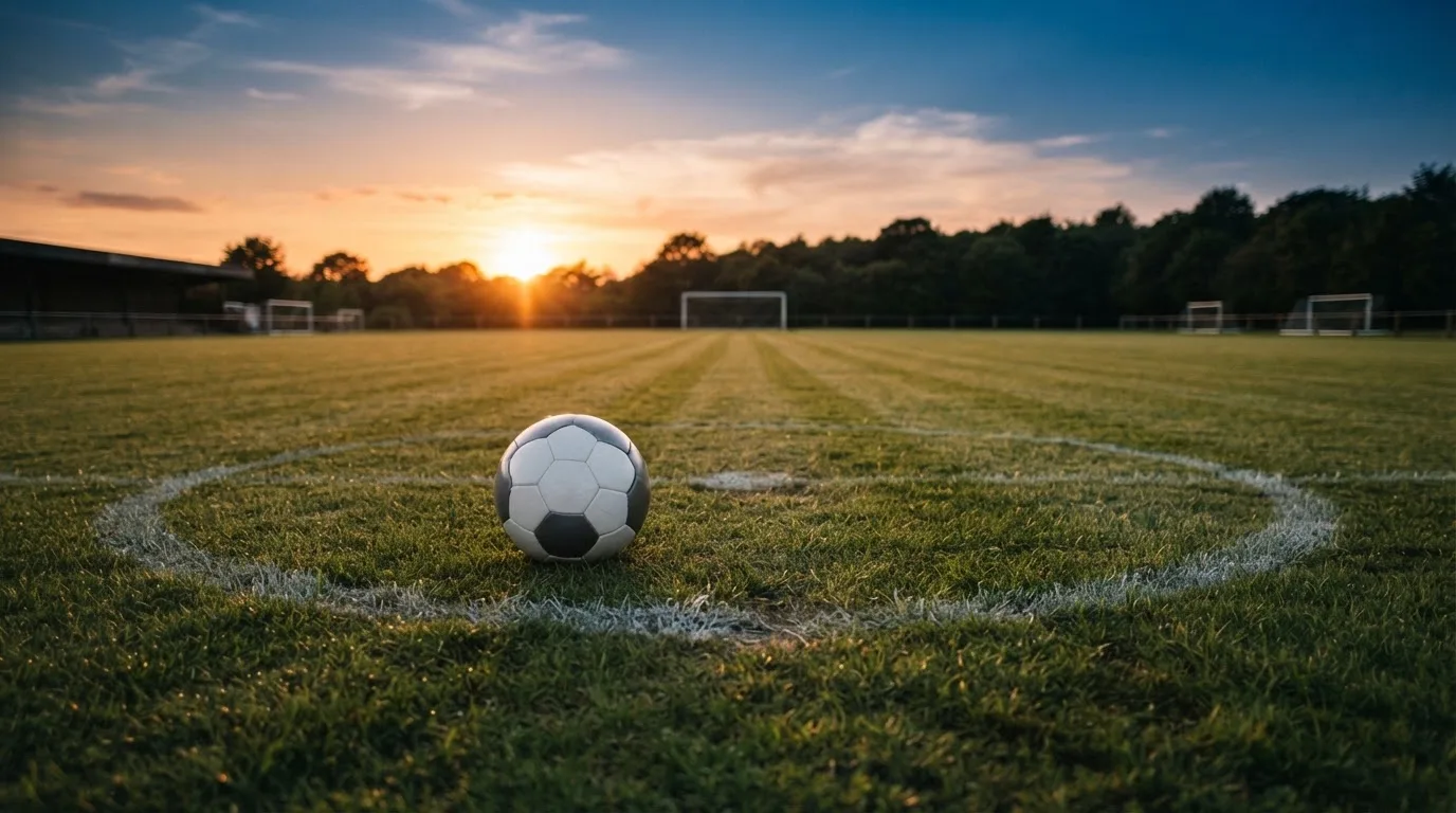 Ein Fußball liegt in der Mitte eines leeren Rasenplatzes, am Horizont färbt sich der Himmel orange.