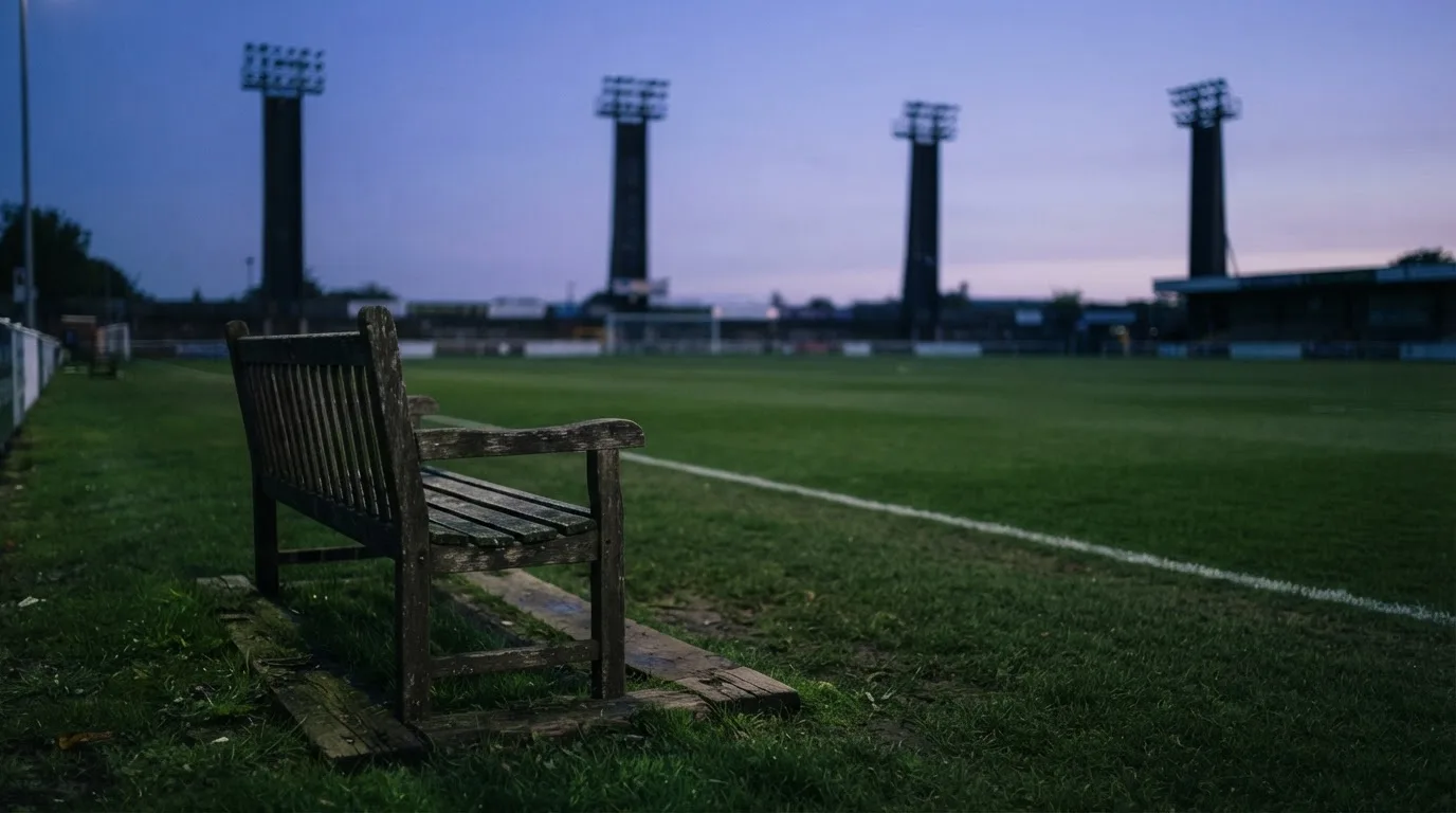 Eine leere Parkbank am Rand eines Fußballfelds in der Abenddämmerung, im Hintergrund ragen dunkle Flutlichtmasten in den Himmel.