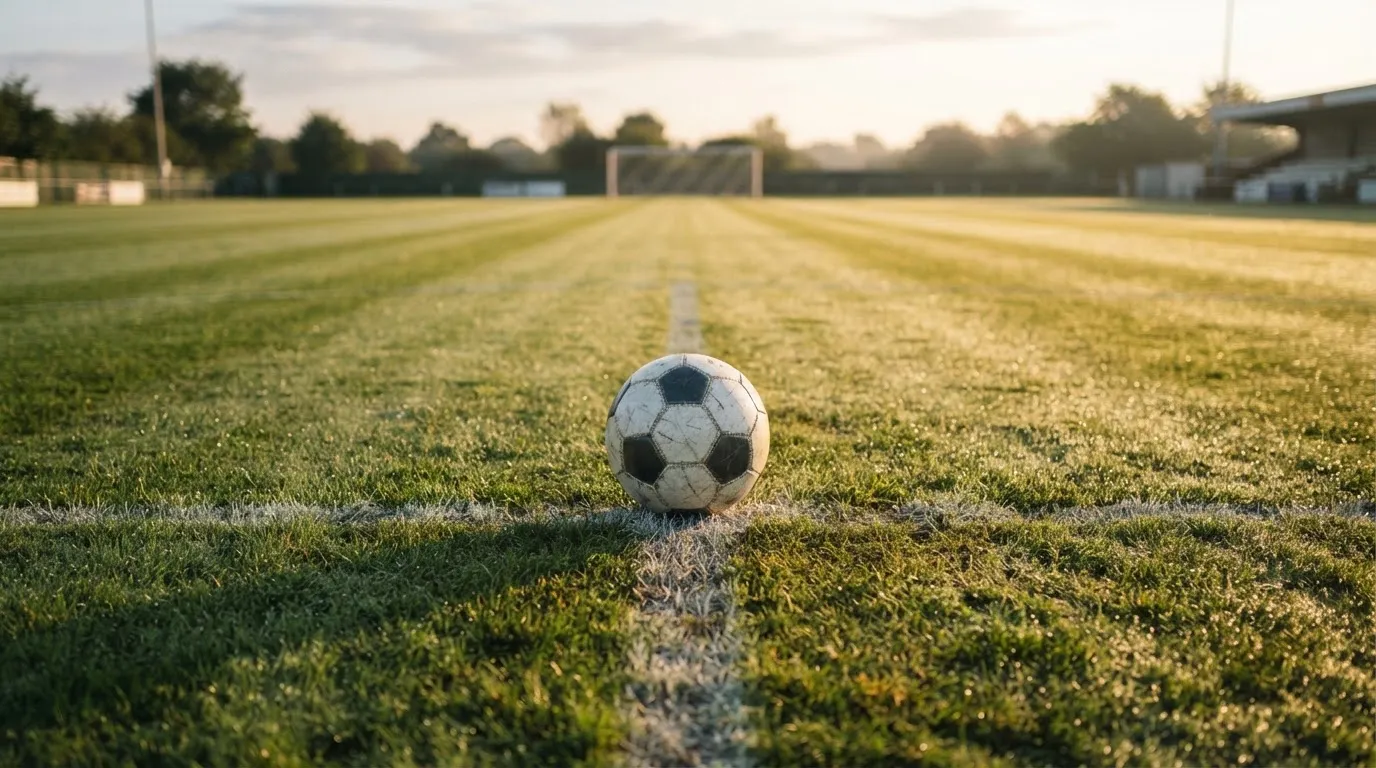 Ein Fußball liegt auf dem Anstoßkreis eines leeren Rasenplatzes im Licht der Morgensonne.