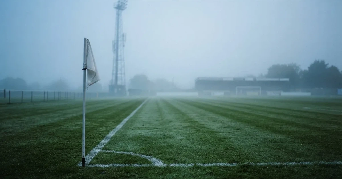 Eine Eckfahne steht im Nebel am Rand eines leeren Rasenplatzes in der Morgendämmerung, dahinter ein Flutlichtmast.
