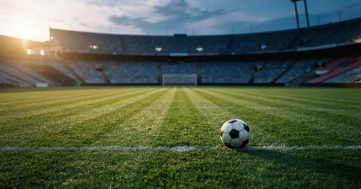 Ein Fußballplatz mit frisch gemähtem Rasen im Abendlicht, ein Ball liegt im Strafraum, im Hintergrund unscharf eine leere Tribüne.