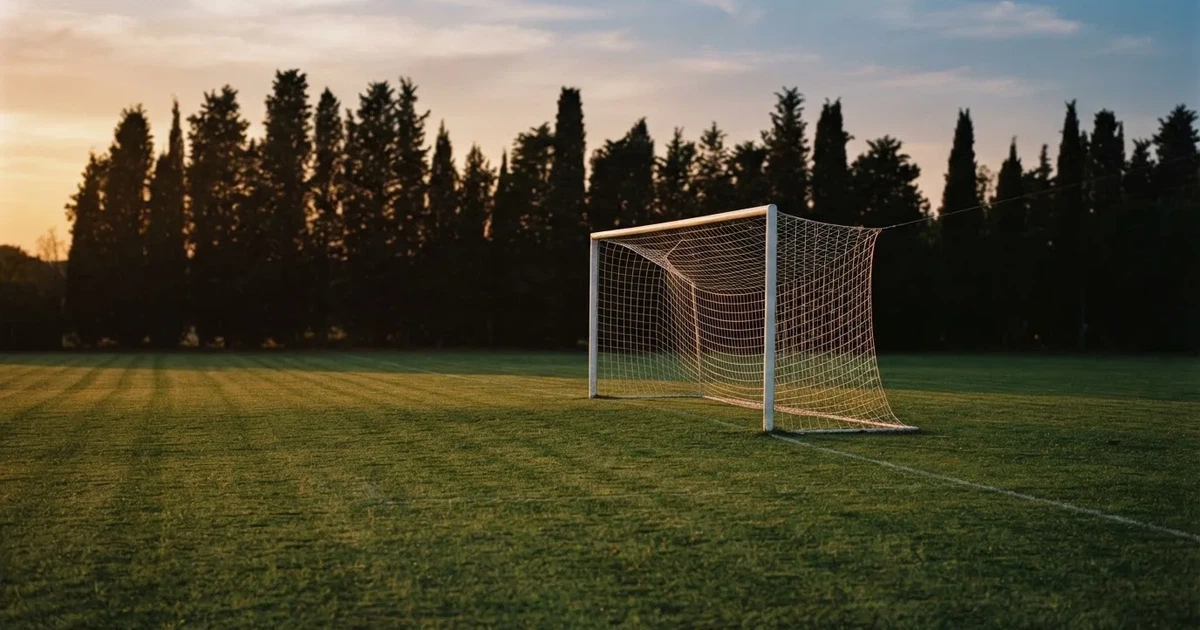 Ein Fußballtor mit Netz auf einem leeren Platz im Abendlicht, der Rasen frisch gemäht, im Hintergrund Zypressen.