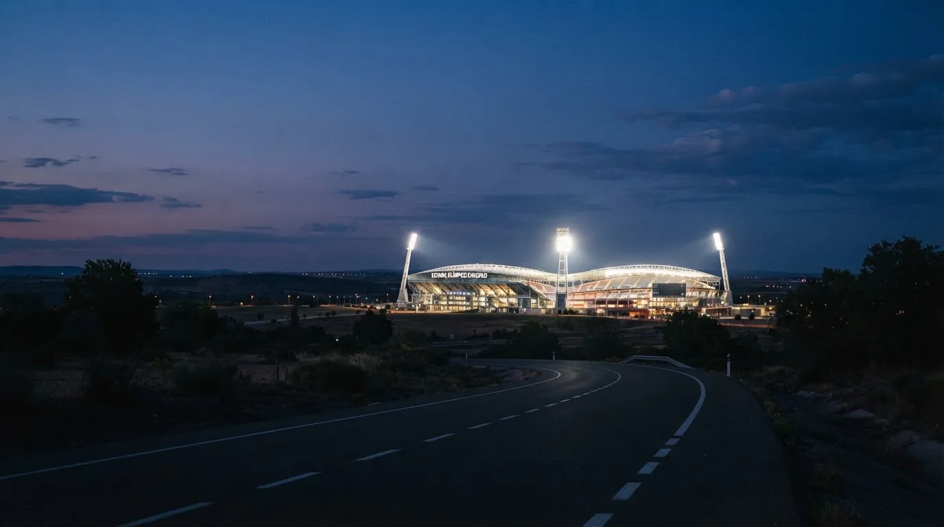 Ein Flutlichtstadion in der Abenddämmerung aus großer Entfernung, die Lichtmasten brennen, davor eine leere Landstraße.