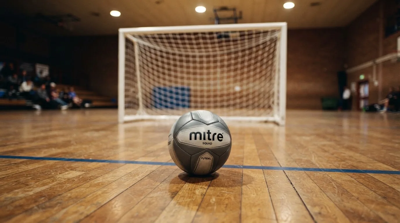 Ein kleiner schwerer Ball liegt in einer Halle auf dem Parkett vor einem Tor, warmes Licht aus den Deckenstrahlern.