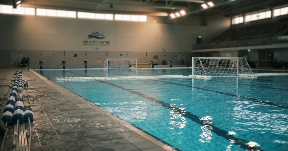 Ein Schwimmbecken mit türkisfarbenem Wasser und zwei schwimmenden Torrahmen am gegenüberliegenden Beckenrand, menschenleer unter Hallenlicht.