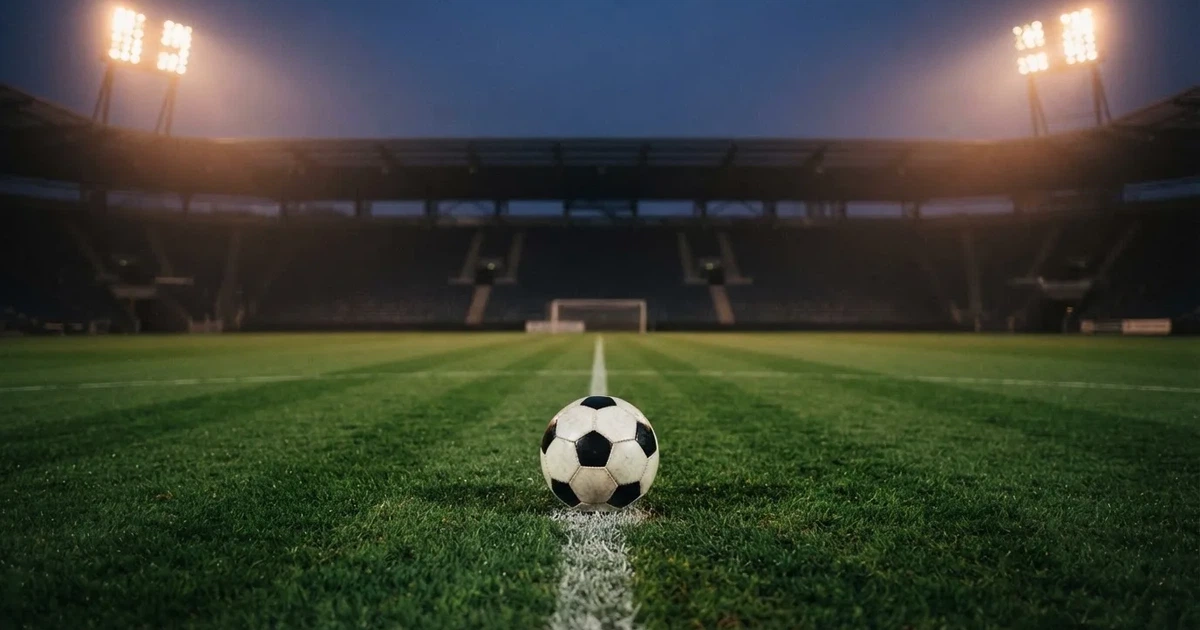 Ein Rasenplatz in der Dämmerung mit eingeschalteten Flutlichtmasten, ein Ball liegt am Anstoßpunkt, die Tribünen im Hintergrund sind unscharf.
