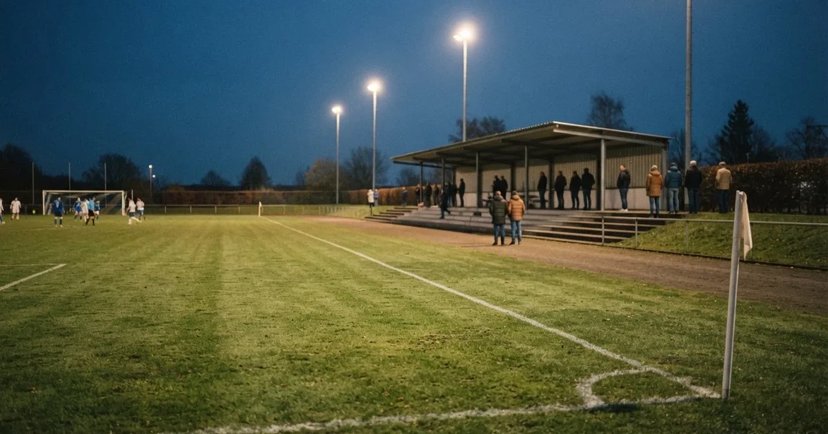 Ein einfacher Rasenplatz mit kleiner Stehtribüne und Flutlichtmasten in der Dämmerung, auf der Tribüne sitzen nur wenige Zuschauer.