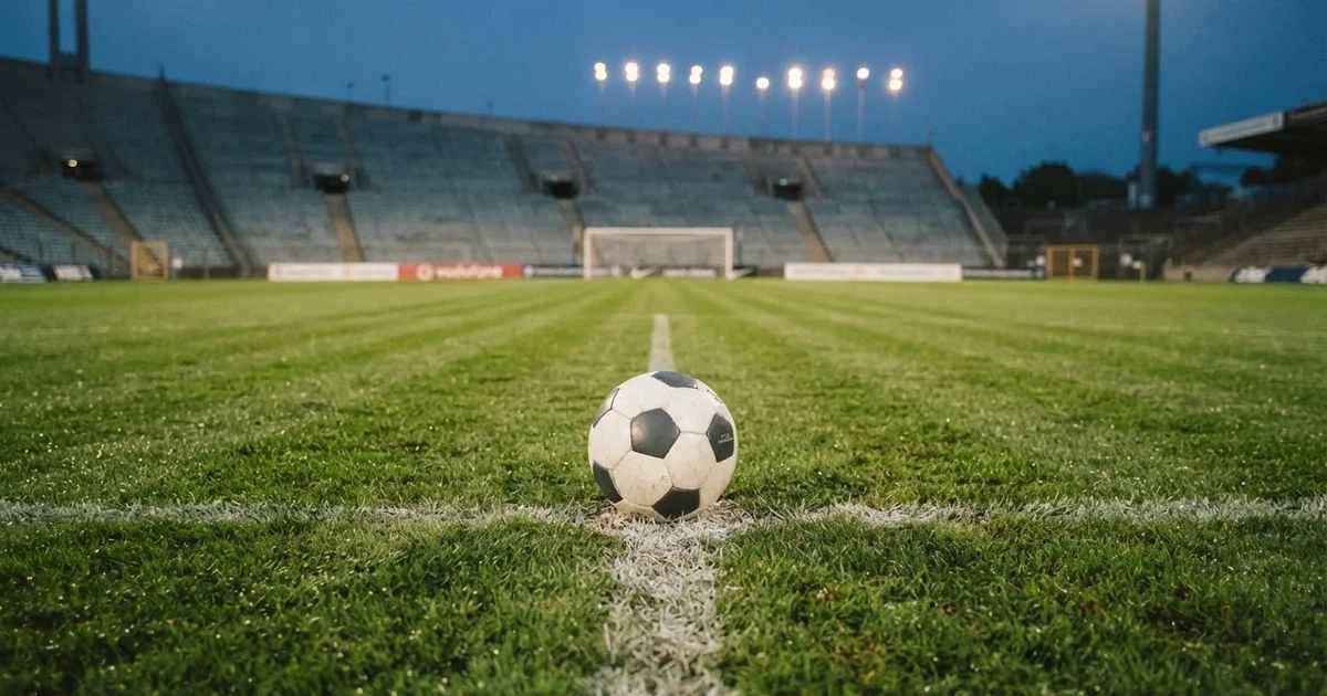 Ein Fußball liegt auf dem Anstoßpunkt eines leeren Rasenplatzes, im Hintergrund eine Tribüne und Flutlichtmasten in der Abenddämmerung.