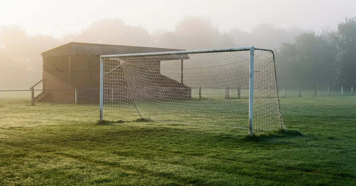 Ein Fußballtor mit Netz steht im Morgennebel auf einem leeren Rasenplatz, im Hintergrund die Silhouette einer kleinen Tribüne.