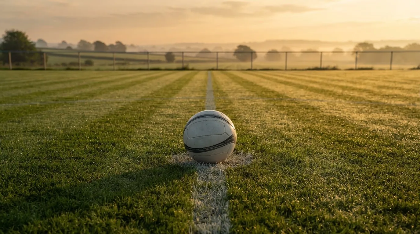 Ein Fußball liegt allein auf dem Anstoßpunkt eines leeren Rasenplatzes im Morgenlicht, am Rand ein niedriger Zaun.