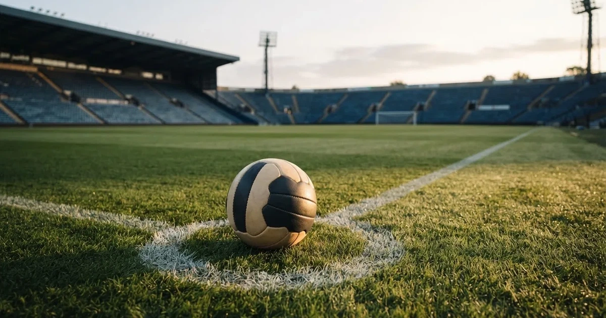 Ein Fußball liegt auf dem Anstoßpunkt eines Rasenplatzes, dahinter leere Tribünen im Licht des frühen Morgens.