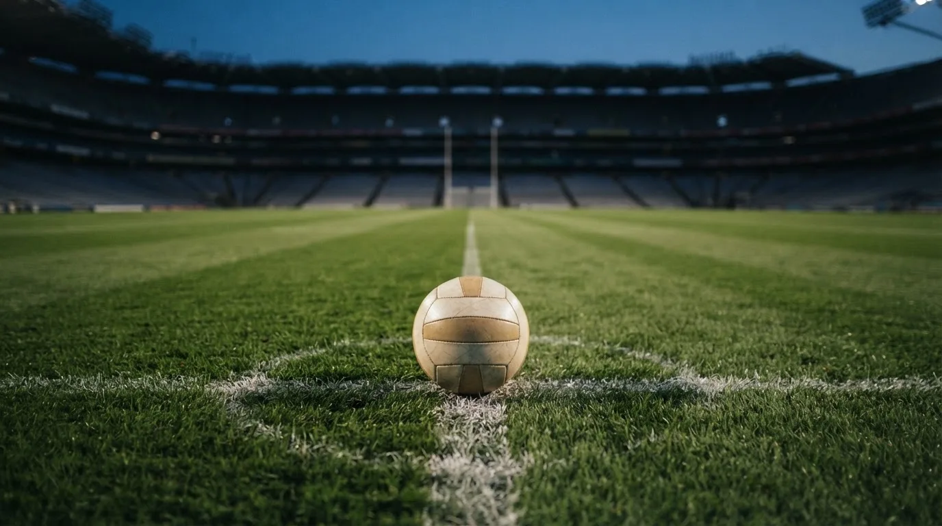 Ein Fußball liegt auf dem Anstoßpunkt eines leeren Rasenplatzes, dahinter ragen dunkle Ränge in den Abendhimmel.