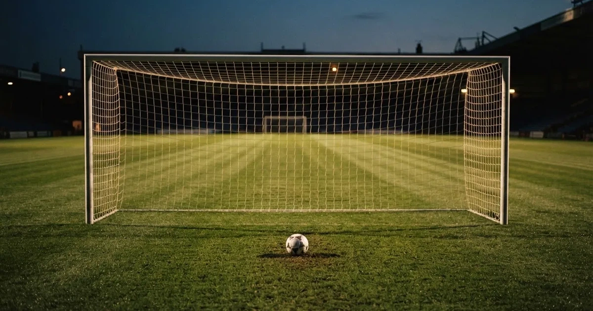 Ein Fußballtor mit Netz im Gegenlicht auf einem leeren Platz am Abend, davor ein Ball auf dem Rasen.