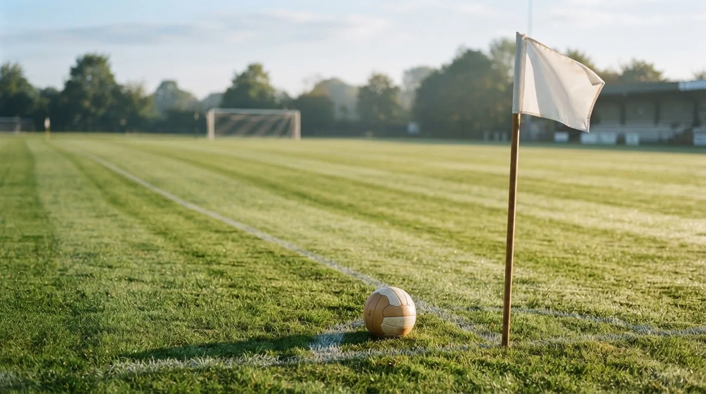 Ein Fußball liegt auf Rasen neben einer Eckfahne ohne Aufdruck auf einem leeren Platz im Morgenlicht.