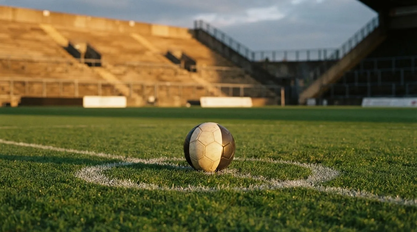 Ein Fußball liegt auf dem Anstoßpunkt eines Rasenplatzes, dahinter eine leere Tribüne im Abendlicht.