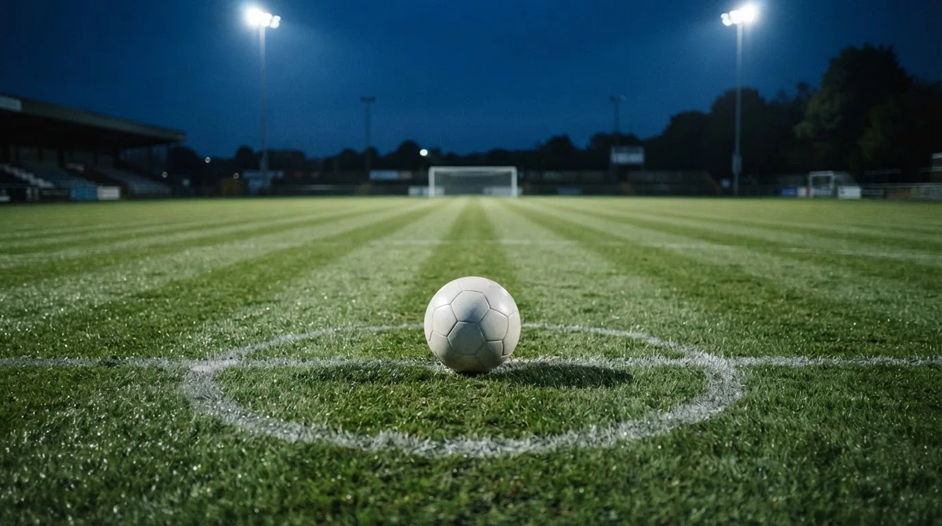 Ein Fußball liegt auf dem Mittelkreis eines leeren Rasenplatzes unter Flutlicht am frühen Abend, die Tribünen sind nur schemenhaft zu erkennen.