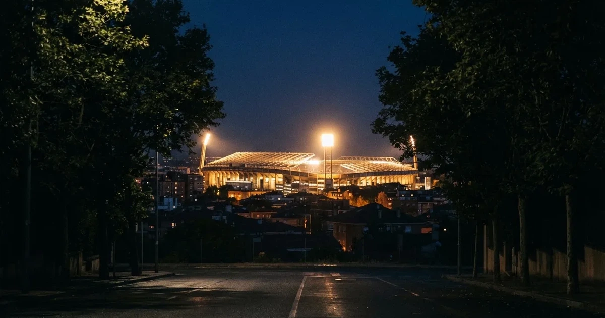 Ein Fußballstadion bei Nacht aus großer Entfernung, hell erleuchtet unter Flutlicht, davor eine dunkle Straße und Bäume.