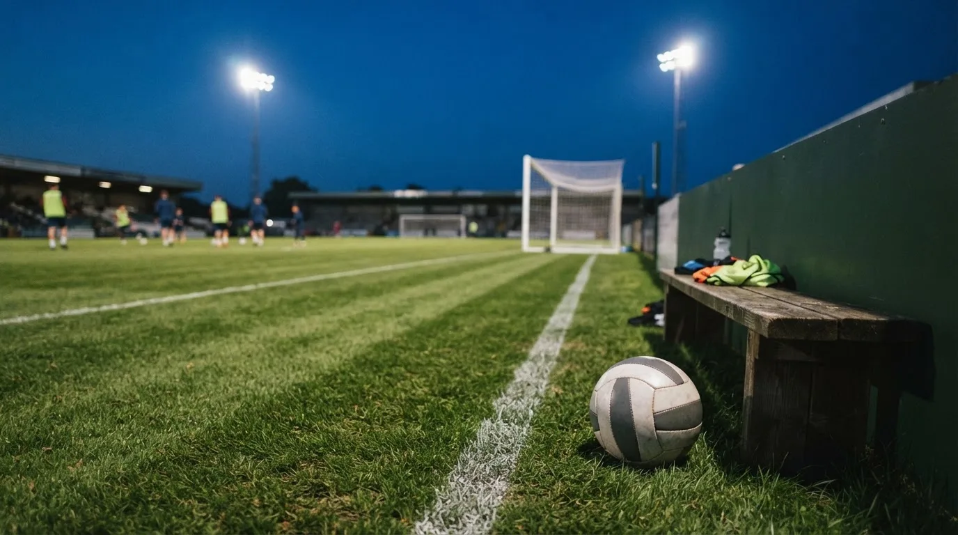 Ein Fußball liegt am Spielfeldrand neben einer leeren Ersatzbank, das Flutlicht über dem Platz leuchtet in der Dämmerung.