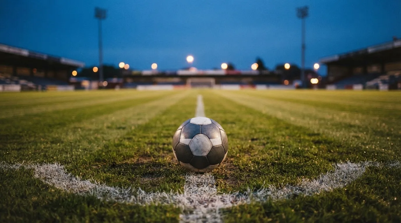 Ein Fußball liegt auf dem Anstoßpunkt eines Rasenplatzes in der Abenddämmerung, Flutlichtmasten stehen unscharf im Hintergrund.