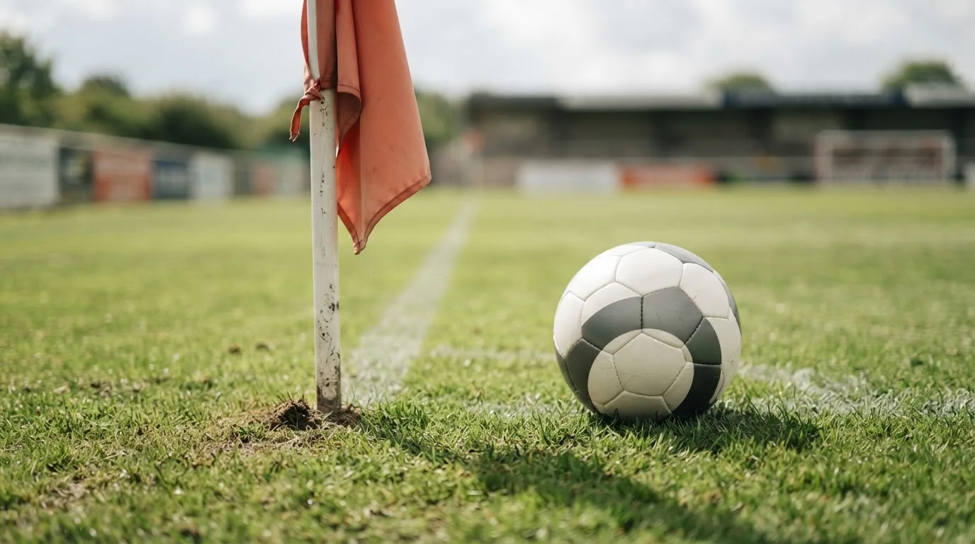 Ein Fußball liegt neben einer Eckfahne auf kurz gemähtem Rasen unter bewölktem Himmel.