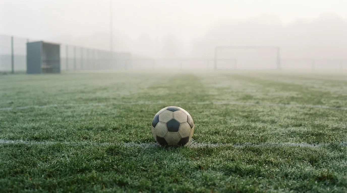 Ein Fußball liegt in der Mitte eines leeren Rasenplatzes im Morgennebel, am Rand steht eine einzelne Auswechselbank.